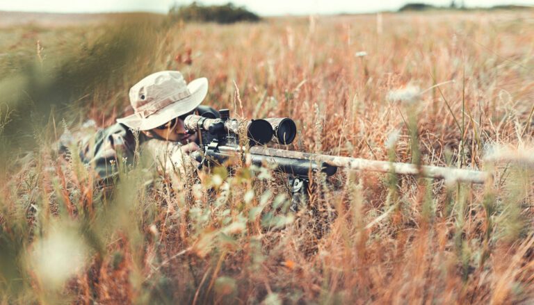 hunter in brown jacket and black pants sitting on brown grass field during daytime