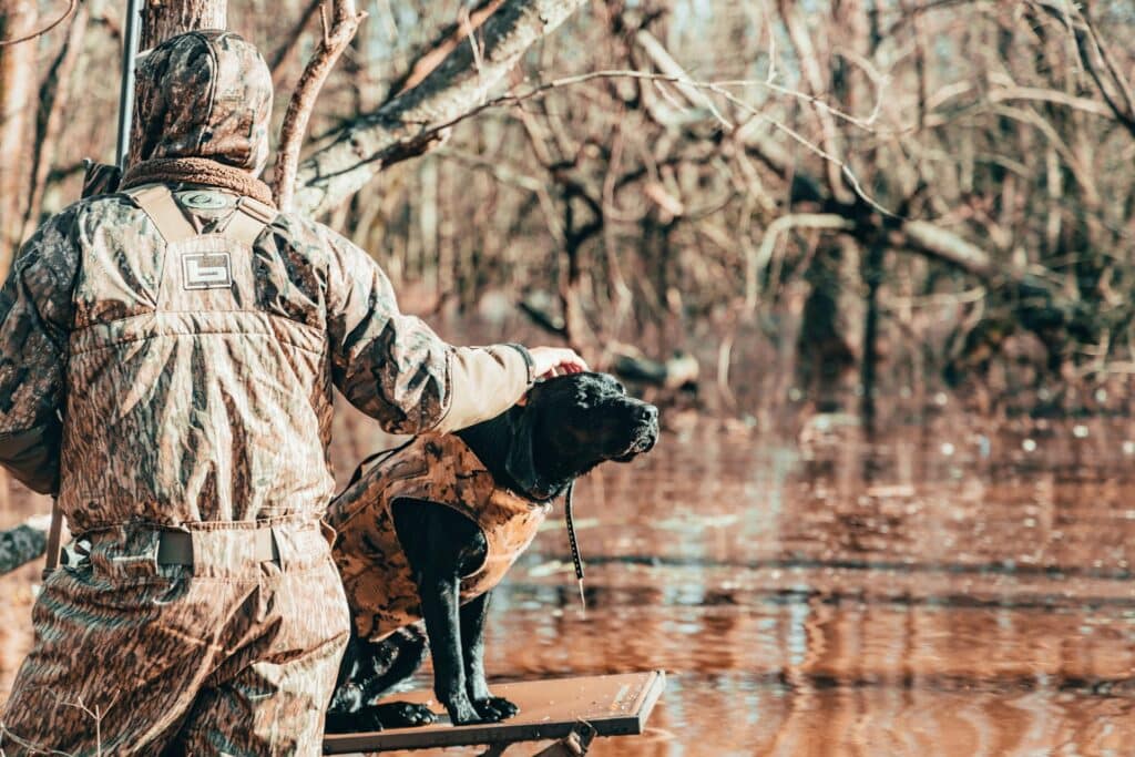 man in hunting gear with his black hunting dog by a lake