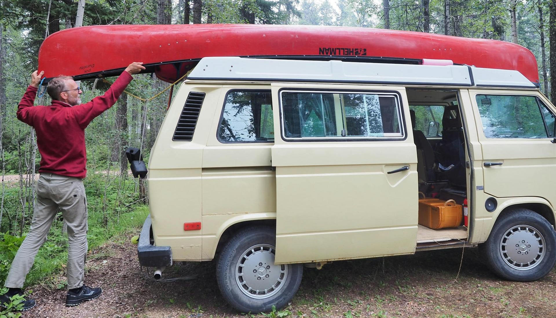 man in red long sleeved shirt putting red canoe on top of off-white van in the woods