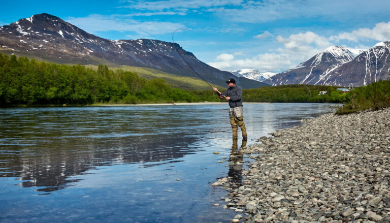 man in a blue jacket fishing on the river with the mountains in the background