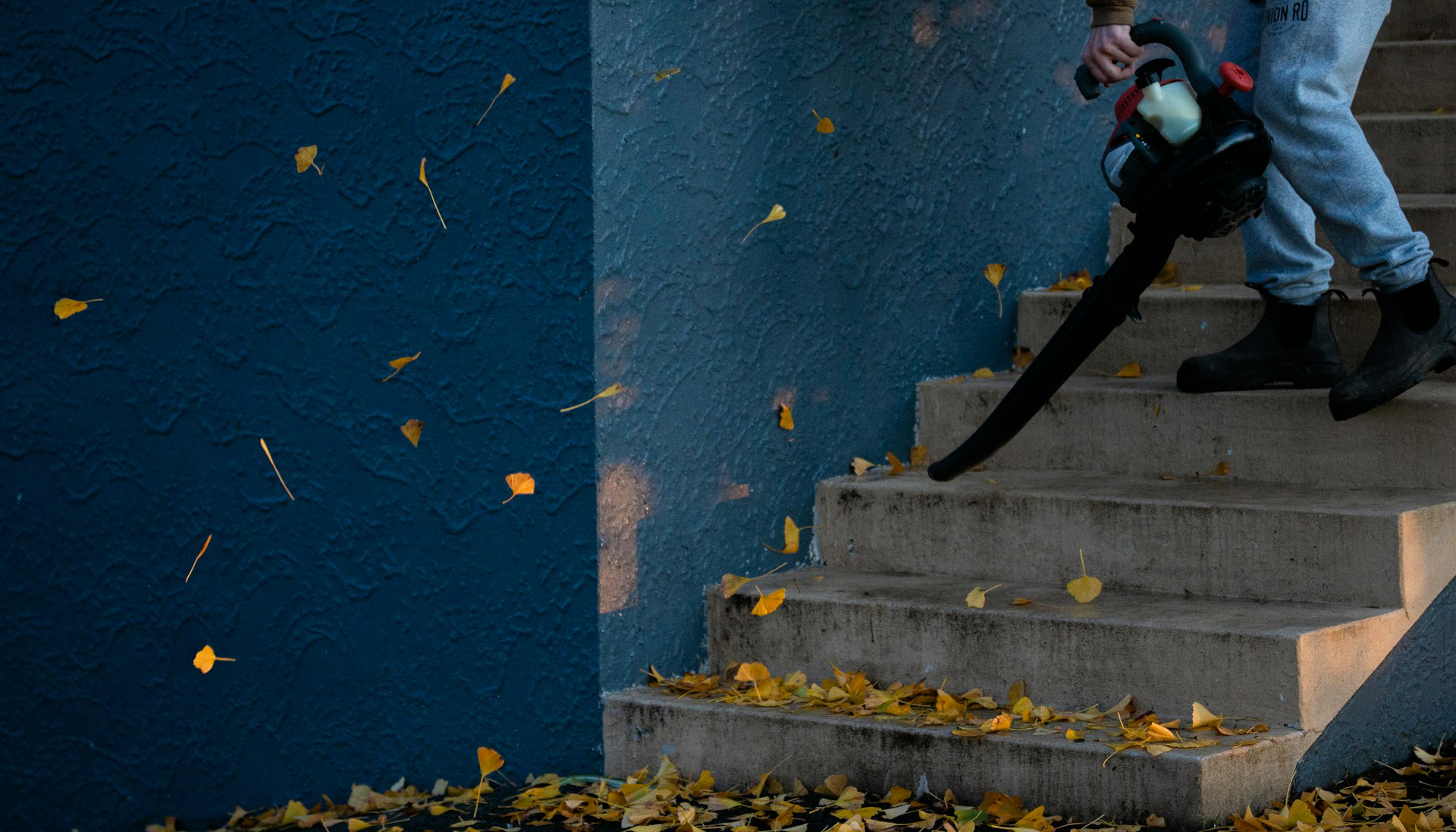 man in black jacket and pants standing on stairs stairs blowing fall leaves using a leaf blower